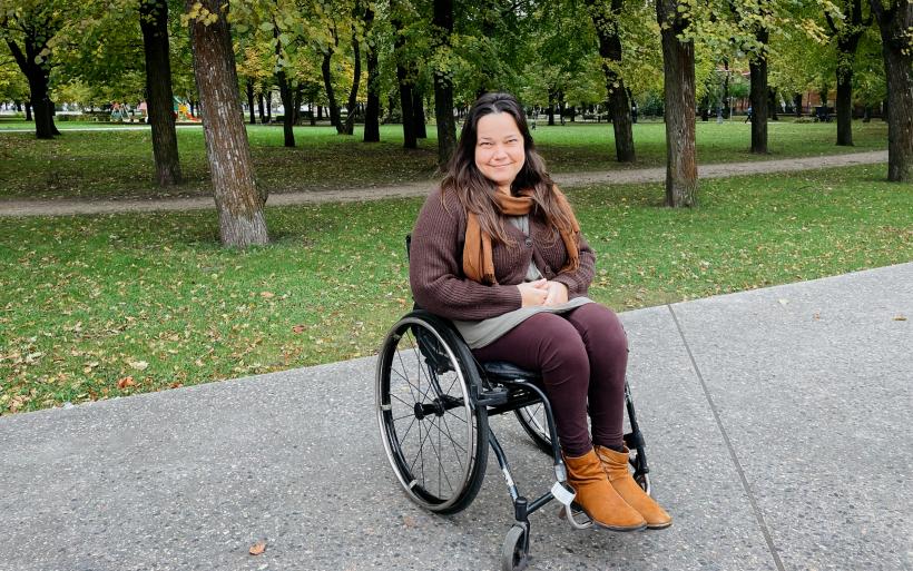 Meitene ratiņkrēslā, fonā parks ar zaļiem kokiem