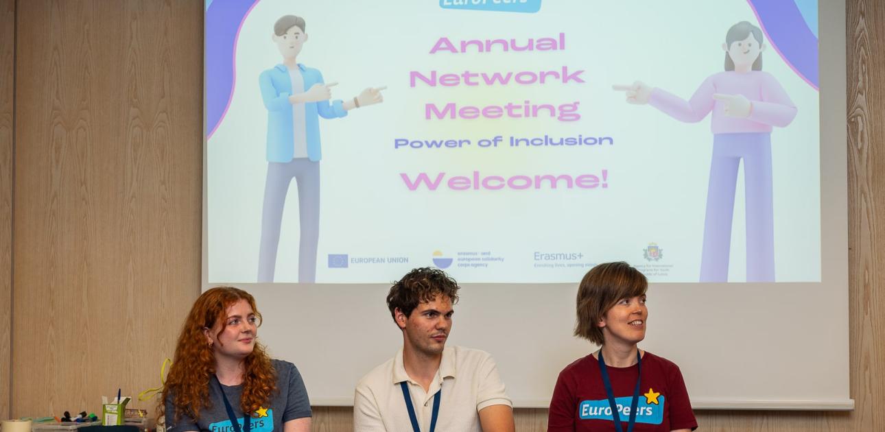 Trīs cilvēki sēž, fonā viņiem prezentācācija ar tekstu - Annual Network Meeting. Welcome!