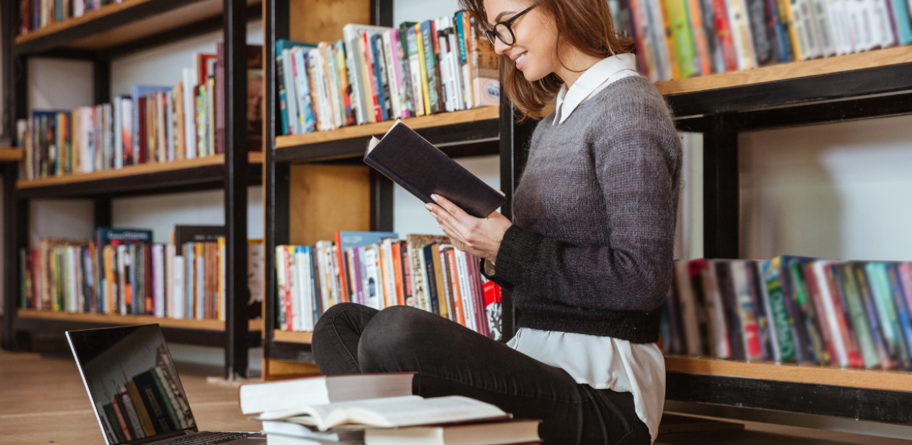 Meitene bibliotēkā sēž uz grīdas un lasa grāmatu