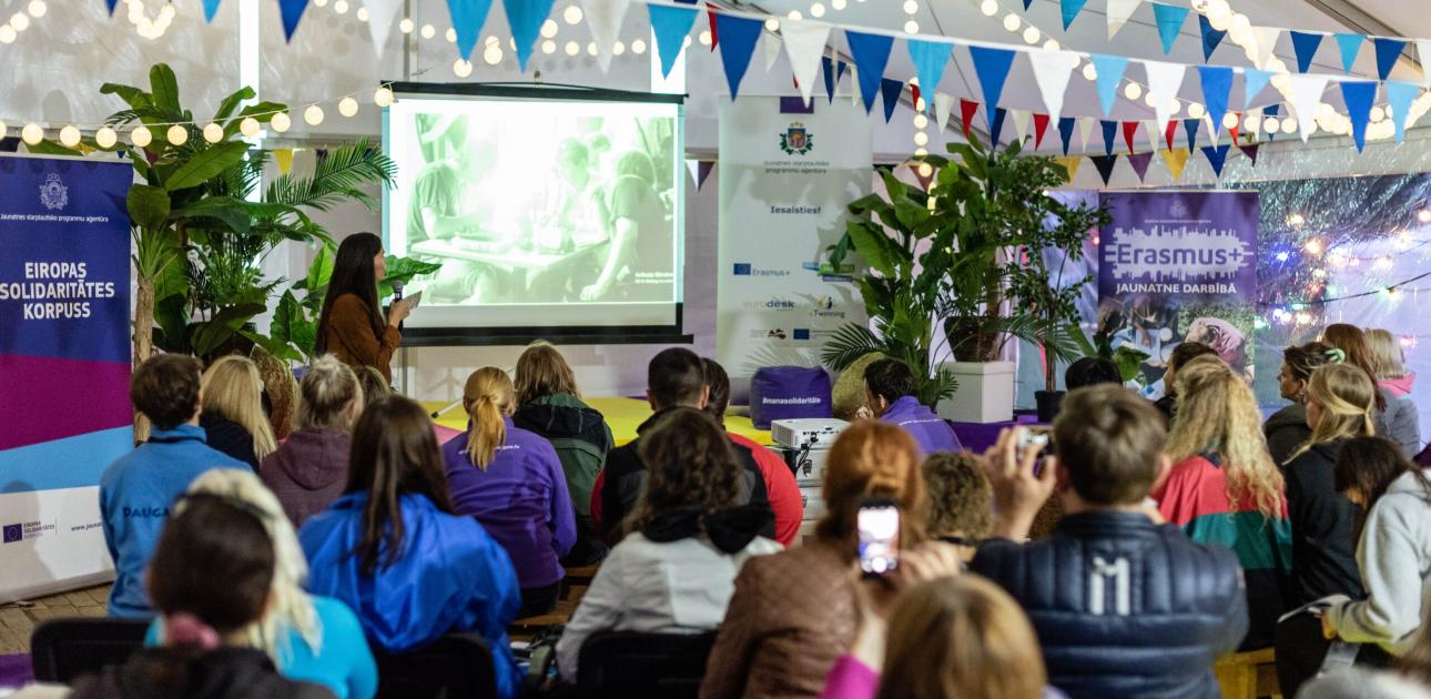 Attēlā no Projektu nakts 2019 redzami cilvēki, kas festivāla noskaņās izdekorētā teltī klausās mācību vadītāja lekciju