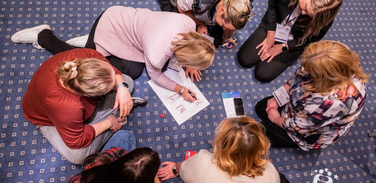 Septiņas sievietes sēž aplī uz paklāja, viena raksta uz lapas