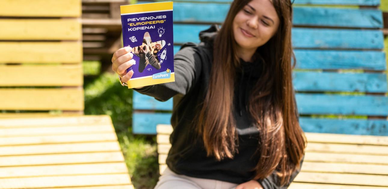 Meitene sēž uz krāsainām paletēm un rokās tur bukletu