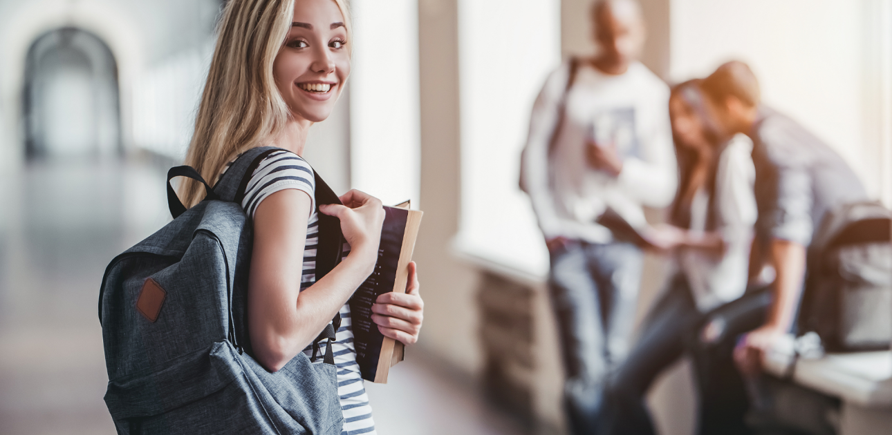 smaidīga studente iet pa universita'tes koridoru ar mugursomu un grāmatām rokās, aizmugurējā plānā citi studenti