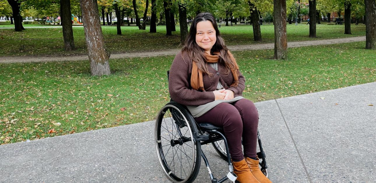 Meitene ratiņkrēslā, fonā parks ar zaļiem kokiem