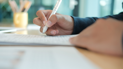 attēlā redzama roka ar pildspalvu kaut ko raksta dokumentā