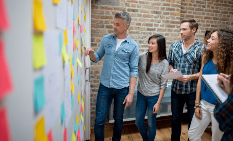 Četri cilvēki stāv pie tāfeles un līmē līmlapiņas uz tās