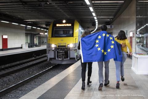 Jaunieši apsegušies ar Eiropas Savienības karogu iet pa dzelzceļa staciju
