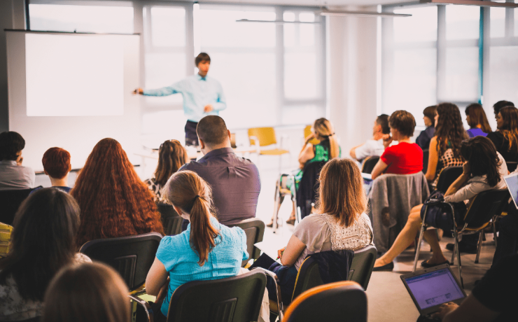 Attēlā redzami cilvēki, kas sēž un klausās runātājā
